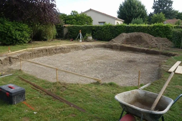 Tranchée de piscine prête pour une dalle, avec piquetage et remblais, jardin entouré d’arbres.