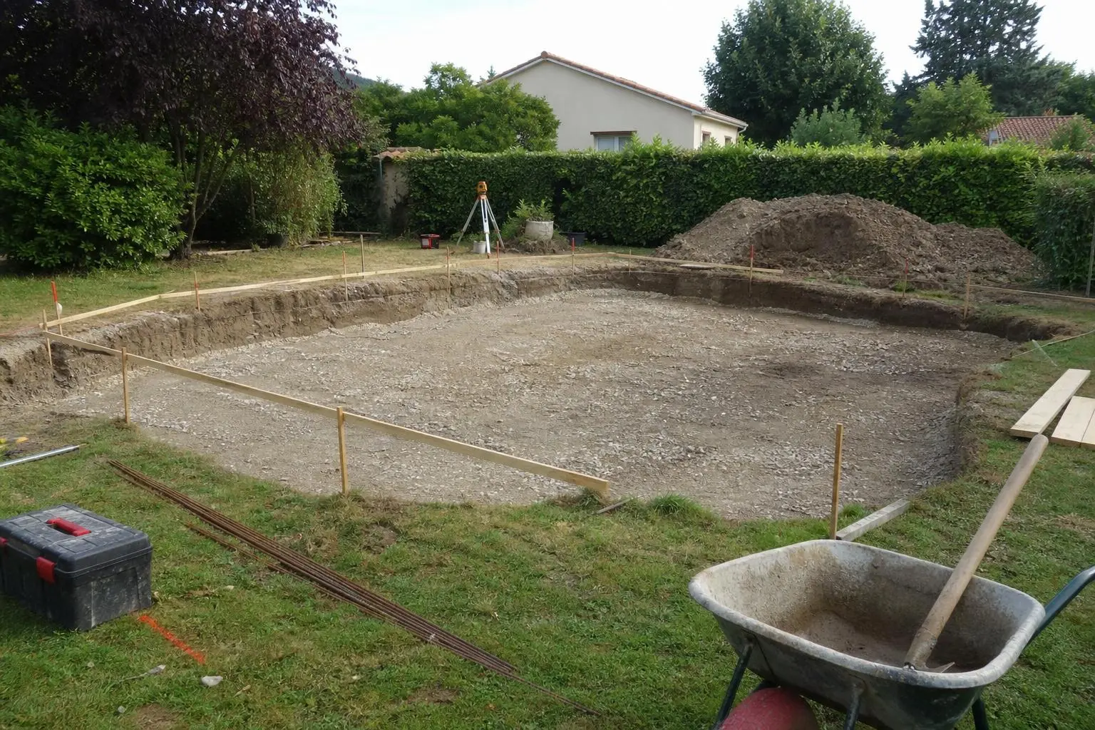Tranchée de piscine prête pour une dalle, avec piquetage et remblais, jardin entouré d’arbres.