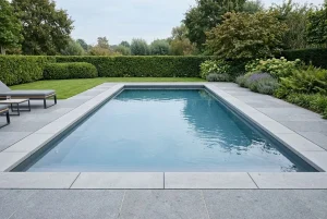Piscine enterrée moderne en retrait de terrasse pavée, entourée de haies et de verdure, avec mobilier extérieur. piscine enterrée combien coûte latent dans l’image.